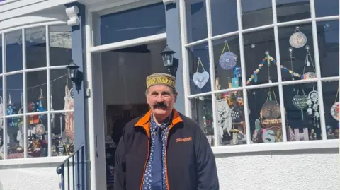 John Starkey Clairvoyant A man in a blue coat with an orange trim, is wearing a hat and has a thick handlebar mustache, He is standing in front of a shop with trinkets in the windows