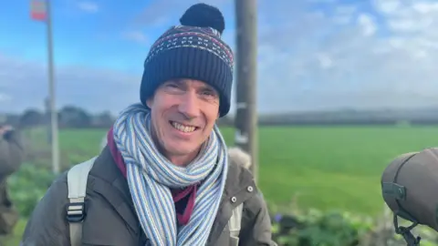 Man wearing a patterned bobble hat and stripey scarf, standing beside a camera on a tripod. he is smiling with fields behind