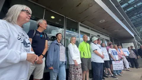Campaigners, all wearing a range of different outfits and some holding placards which state 'stop vandalising culture and heritage' and 'Save the National Glass Centre' stand in front of the now permanently closed building. They are in front of large glass-fronted doors, which have signs in their windows stating there is security now operating.