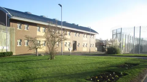 A long two-storey building in light brown and yellow brick. There is a grassy area to the front and a fenced area to the right with barbed wire along the top.