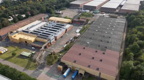 Michelin An aerial view of a factory site, with warehousing stretching into the distance on the right of the picture, and trees on either side of the site.