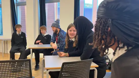 BBC Six secondary school pupils sit around two small, square tables in a classroom alongside a man in Bristol Bears kit and beanie hat. The pupils are a mixture of boys and girls and wear dark school jumpers over white shirts and ties.