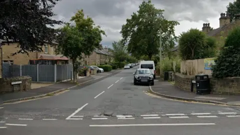 Google A road stretches into the distance, with cars parked on both sides. It is a residential street and homes with driveways and fences can be seen on both sides of the road. Trees line the street.