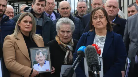 PA Media Three women holding photographs of Sean Brown standing at microphones outside. A group of about 10 men are standing behind the.