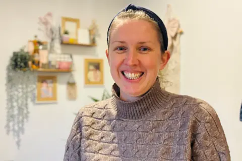 A photo of Stacey Barr smiling at the camera, she is wearing a beige knitted jumper.