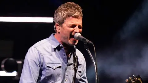 PA Media Noel Gallagher in blue denim sings into a microphone at the Oasis comeback gig in Cardiff