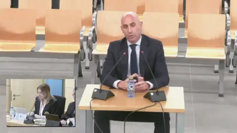 Reuters Rubiales, with a suit and tie, sitting at a table while evidence in a court room. 