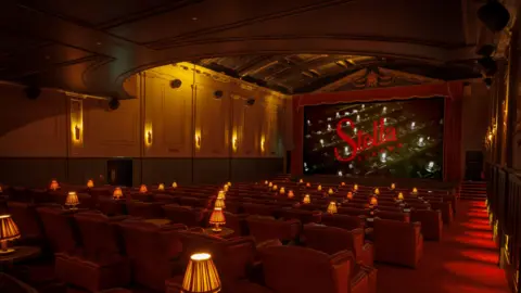 Stella Cinema A cinema with red velvet chairs facing a screen that says Stella Cinema. Lamps have been placed throughout the cinema. 