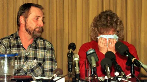 PA Media Paul and Janet Betts sitting in front of microphones lined up on a desk at a press conference. Janet is covering her face with a tissue while crying, head in hands. Paul is also visibly emotional while looking at his wife.