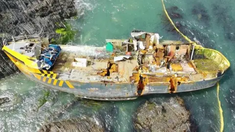 A shipwreck is seen from a bird's eye view. It sits on rocks and is tilted at a slight angle. The ship has two gashes in its side.