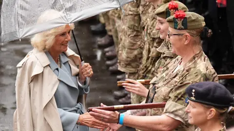 PA Media Queen Camilla wearing a blue dress and beige trench coat holding an umbrella shakes the hand of a female soldier in a camouflage uniform.