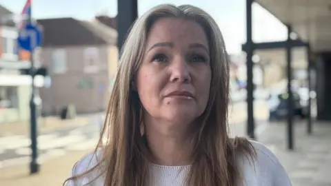 BBC/Simon Thake A woman with long fair hair and wearing a white jumper stands in a town centre looking dejected 