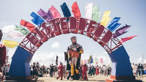 Aled Llywelyn | Eisteddfod Genedlaethol The puppet of Lord Rhys ap Gruffyd is guided through a giant blue arch, with Eisteddfod written in large letters upon it. There are different coloured flags flying from the arch and a crowd of people watching the procession.