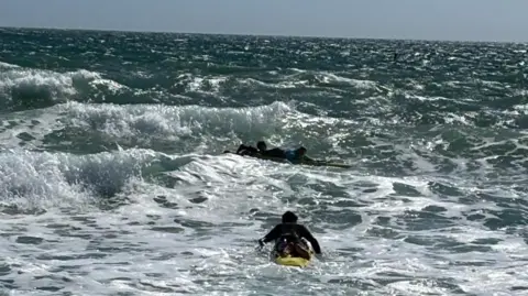 One of the surf lifeguards is on a paddleboard paddling out to two young people on surfboards in choppy water on a sunny day.