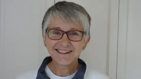 Give a Kidney Sarah Lovegrove smiles at the camera, wearing glasses and a white jumper with a blue collar