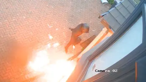 Hassan Patel A man dressed all in black pours fluid on a fire at the front of a house.