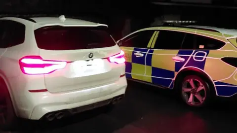  A white BMW next to a police car. The picture shows the back of those cars as they are parked at night.