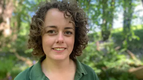 A woman with short curly hair is looking at the camera. She is wearing a green t-shirt and she is standing in a wooded area with bluebells in the background.