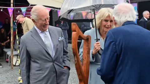 Pacemaker The king is smiling widely while admiring a wooden sculpture of a harp, the Queen is holding a see-through umbrella and is talking to an older, white-haired man who has his back to the camera. 