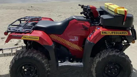 BBC A red and black quad bike. It has RNLI branding on it and small black wheels. It is parked on a beach.