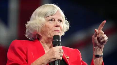 Getty Images Ann Widdecombe speaking at the Reform UK party conference in 2024. She has a bob style haircut with mostly white hair, is wearing a red blazer with a broach, and is holding a microphone in her right hand as she speaks, while making a pointing gesture with her left hand.
