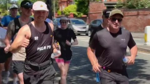 Two men are prominent at the front, with both wearing caps and black clothing. Other runners are behind them. The man at the front on the left is acknowledging being filmed and the group is about to pass the camera. 