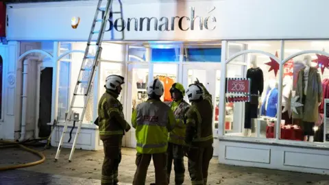 Four firefighters outside a white shop with the name Bonmarche above the door at night