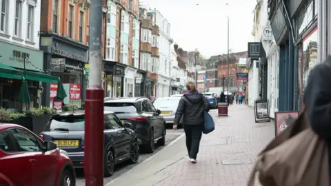 High Street in Tunbridge Wells, lined with shops. The road is busy with parked cars and there is a lone pedestrian walking on the pavement.