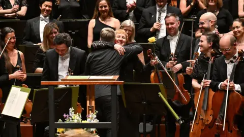 Mark Allan Jacoba Gale and Kirill Karabits embracing at her final concert in Poole while the rest of the orchestra look on