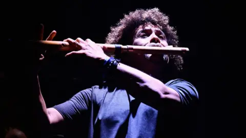 Getty Images A man with curly, afro-style hair plays a modified woodwind instrument that resembles a flute. He's holding one end up to his mouth, and both arms are supporting the other end of the long instrument at chin height. He appears to be standing up, and exerting himself as he plays. He's bathed in a stage light that illuminates the front of his body and face but quickly fades off into blackness behind him.