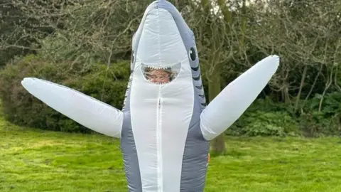 Georgie Box in her shark fancy dress. Georgie is a 28-year-old white woman and is pictured inside an inflated shark costume with both arms held up. The shark's smiling mouth provides a window through which you can just about make out Georgie's blonde hair. She's pictured outside in a park on a bright day