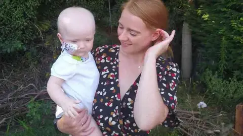 Family Callan Brett, 33, of Ipswich, with her baby son Henry in a woodland setting. Baby Henry has a breathing tube affixed to his nose and wars a white Babygro. Callan is stroking her hair behind her ear and is smiling at her son