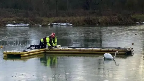 Cambridgeshire Fire and Rescue Large lake with two firefighters crouching on an inflatable path to reach a swan stuck in ice