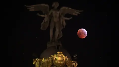 Reuters The moon is seen beside "Victoria Alada" statue on the top of Metropoli building during a total lunar eclipse, known as the "Super Blood Wolf Moon" in Madrid, Spain