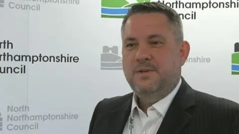 BBC Man with short dark hair and faint beard wearing a jacket in front of North Northamptonshire Council signs