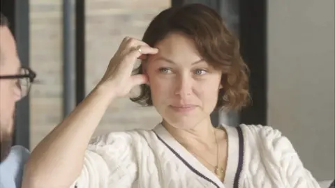 TV presenter Emma Willis, wearing a white jumper and holding her hand to her head