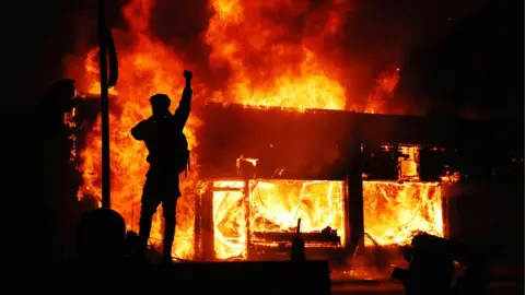 30 May 2020 A protester gestures as buildings burn during continued demonstrations against the death in Minneapolis police custody of African-American man George Floyd, Minnesota