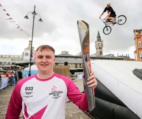 Getty Images Cameron Stewart holds the baton in Darlington as a BMX rider performs a stunt in the background