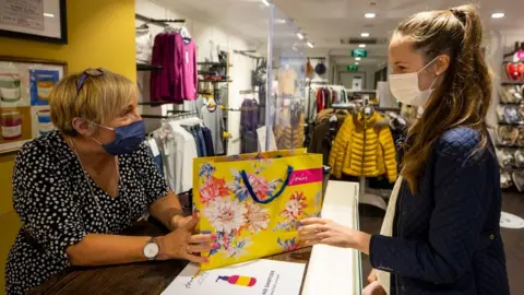 PA Media Mary Rogan, shop assistant at Joules in Belfast, hands Ella Copper her purchase while wearing a face mask as face coverings are now compulsory for shoppers in Northern Ireland