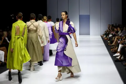 Reuters Models walking on the runway. The model in the middle is wearing a white and purple dress, with the image of a woman on it.