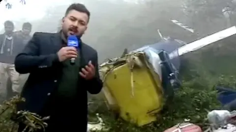 Iranian state TV reporter talking to camera with crashed wreckage of helicopter behind him