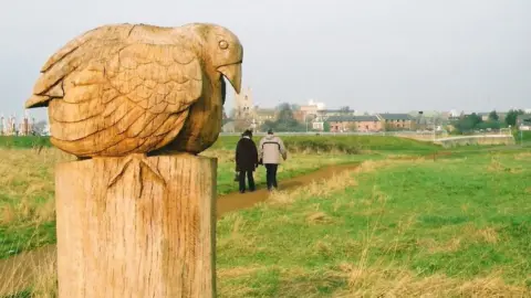 Harding's Pits Community Association View of bird sculpture, two people walking on path across green space and King's Lynn in background