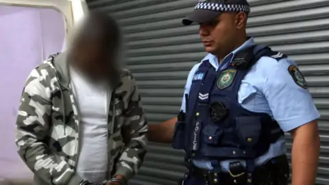 NSW Police The alleged ringleader, shown in an image blurred by New South Wales Police, is accompanied by a police officer