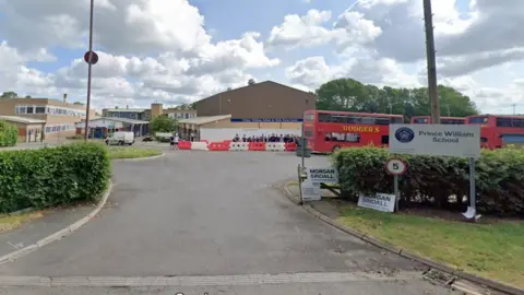 Google Entrance to large school between two hedges. Buses are visible in the background.