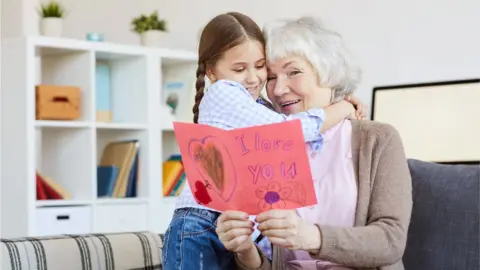 Getty Images Grandmother hugging granddaughter and holding card.