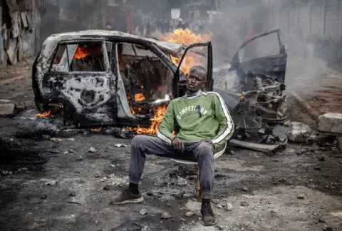 Luis Tato/Getty Images Man sitting down calmly in front of a burning car.