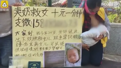 Pear Video Woman kneels on street in China, breastfeeding a baby. The sign beside her has a picture of her ill baby and writing in Chinese characters. There are two plastic bags apparently filled with breast milk in the foreground.