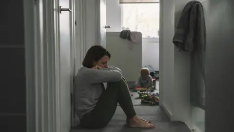 Getty Images Woman looking distressed sits on carpeted floor of home as child plays in background (file image)
