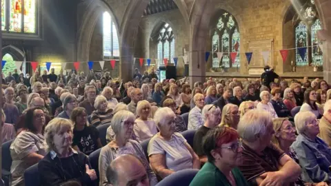 Laura Coffey A church with stained glass windows full of people sitting down for a meeting