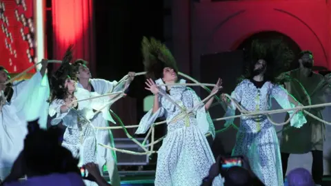 Getty Images A group of performers dancing. Their hair is blowing and they are wearing white dresses.